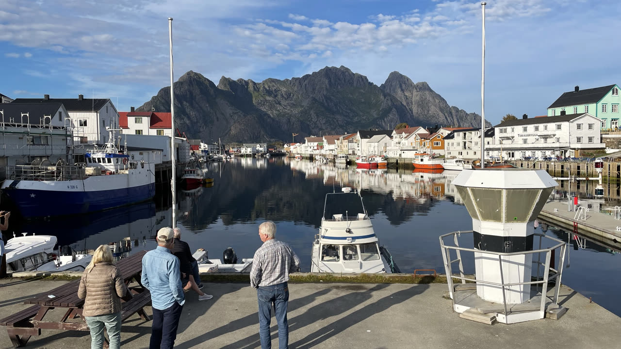 Henningsvær havn i Lofoten en sommerdag, med fiskebåter og hvite hus som speiler seg i stille vann foran snøfrie fjell