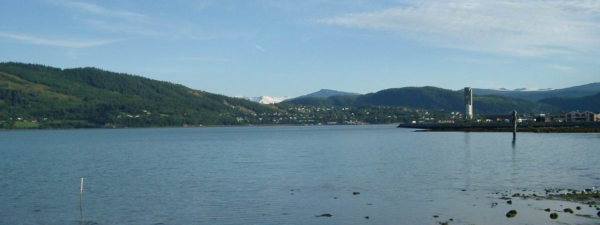 Mo i Rana sett over Ranfjorden med byen langs vannkanten og snødekte fjell i bakgrunnen